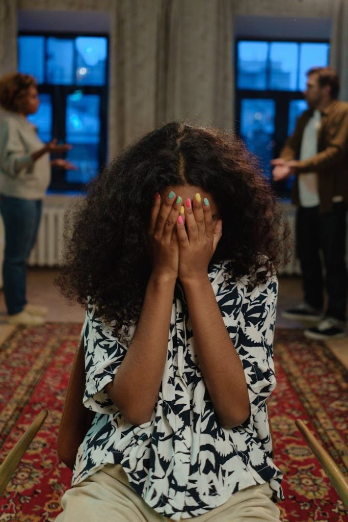 a-girl-in-black-and-white-shirt-covering-her-face-6603345 A young woman sits indoors with her face covered amidst a family argument, conveying distress and conflict.