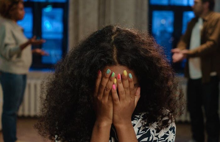 A young woman sits indoors with her face covered amidst a family argument, conveying distress and conflict.