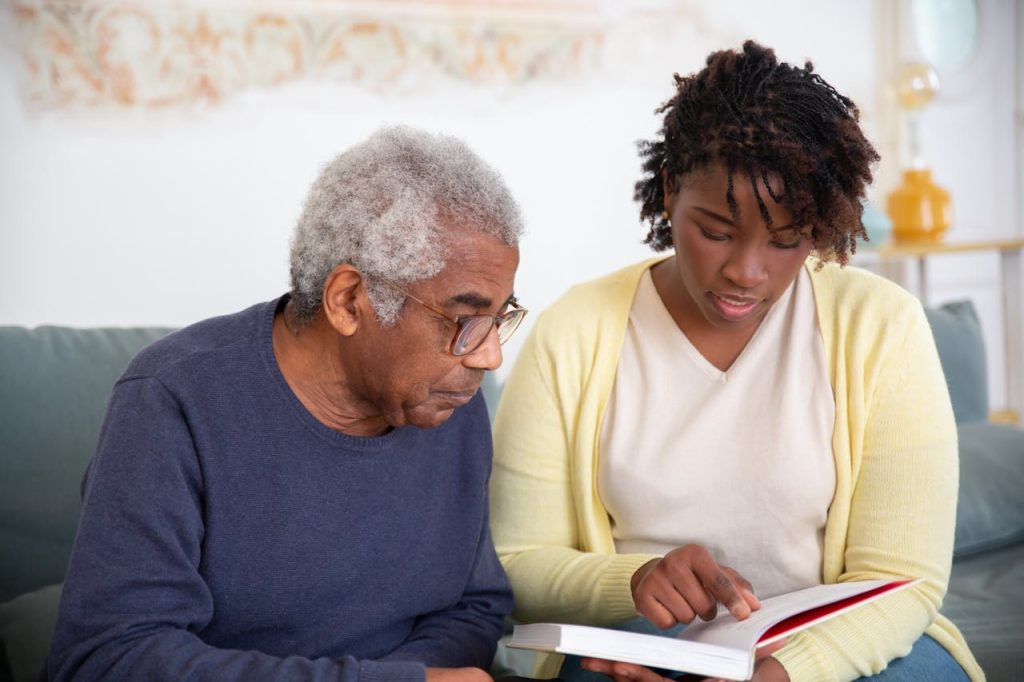 a-woman-in-yellow-cardigan-talking-to-the-man-while-reading-a-book-7551618 A caregiver helps a senior man with reading, providing support at home.